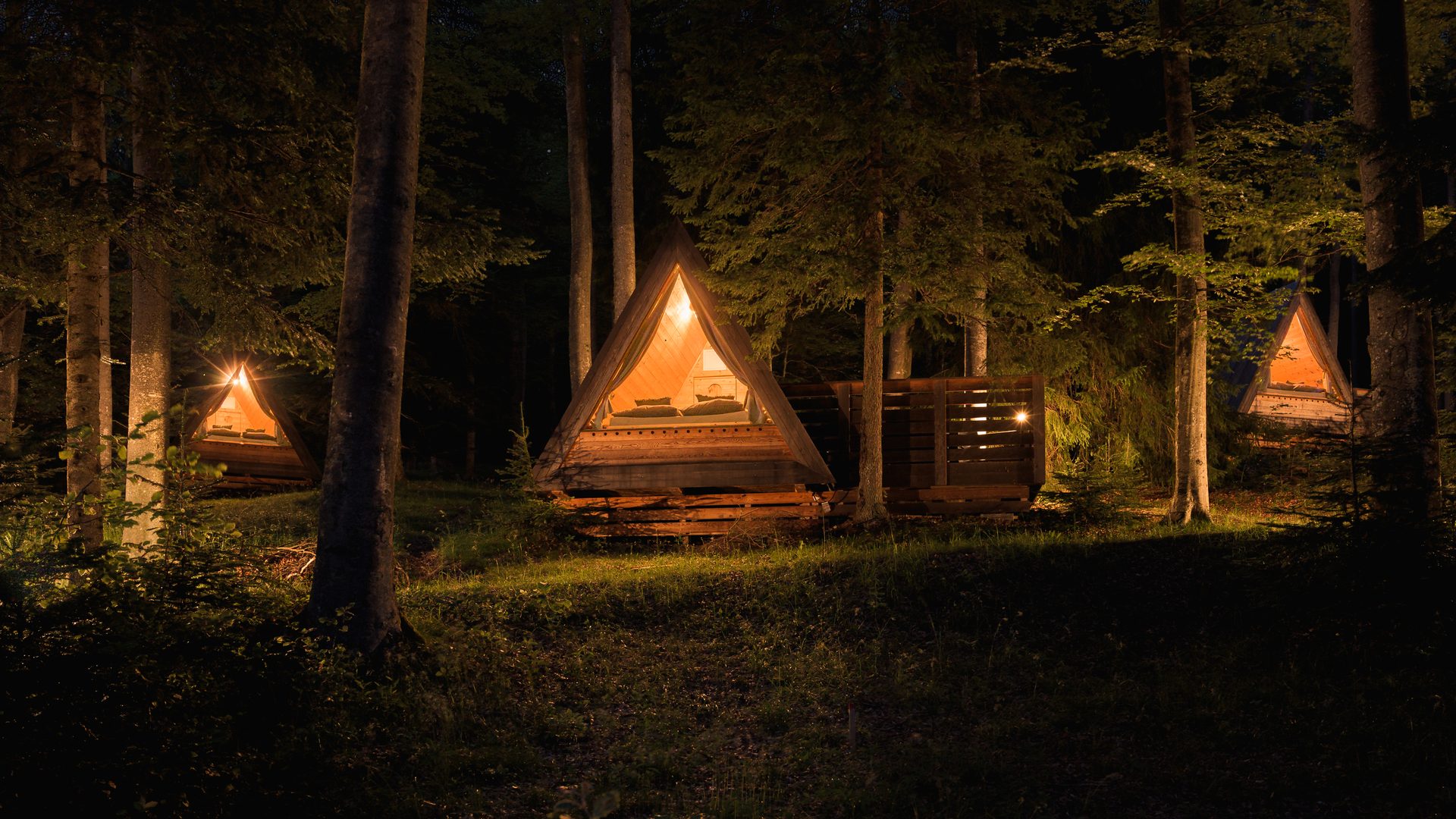 A wooden cabin glowing warmly among tall spruce trees at dusk on Pohorje
